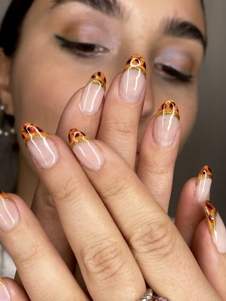 Mano con uñas cortas con punta francesa en tonos naranja y detalles dorados que imitan el efecto de fuego.